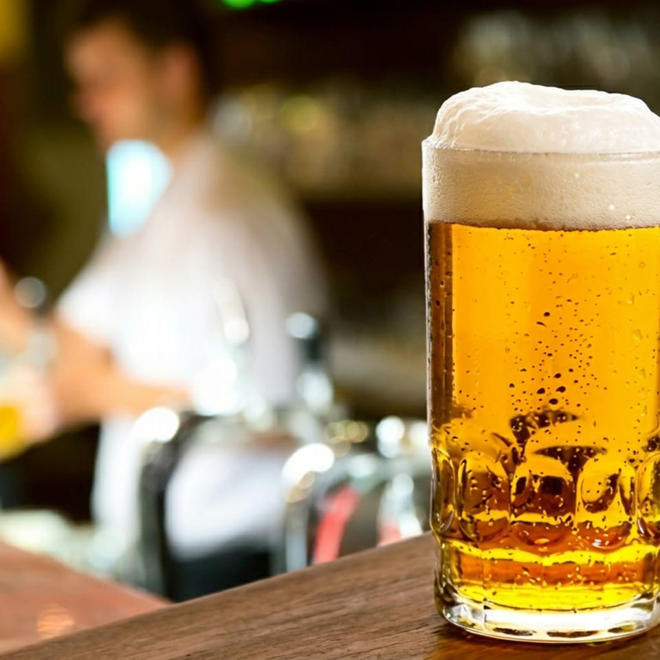 Frosty glass mug of golden beer with a foamy head on a wooden bar, with blurred bartenders and taps behind it.
