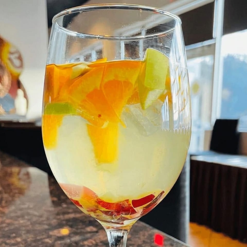 Large glass of pale citrus drink with orange, lemon, lime, and grapefruit slices in a bright restaurant dining area.