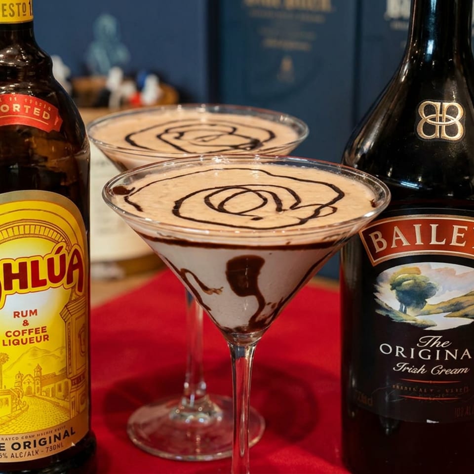 Two creamy espresso martinis with chocolate swirls sit between Kahlúa and Baileys bottles on a red bar counter.