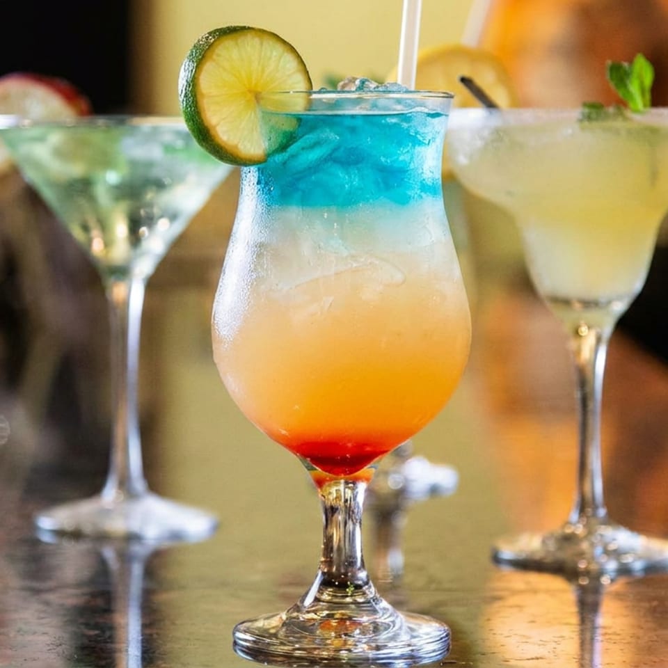 Colorful cocktails, including a layered blue, orange, and red drink garnished with lime, arranged on a restaurant table.