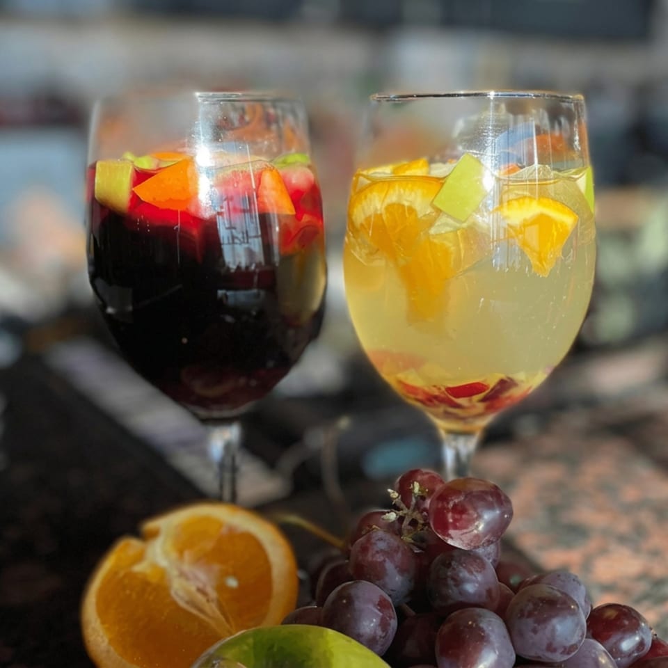 Two stemmed glasses of red and white sangria garnished with fruit, beside grapes, orange, and lime on a restaurant table.