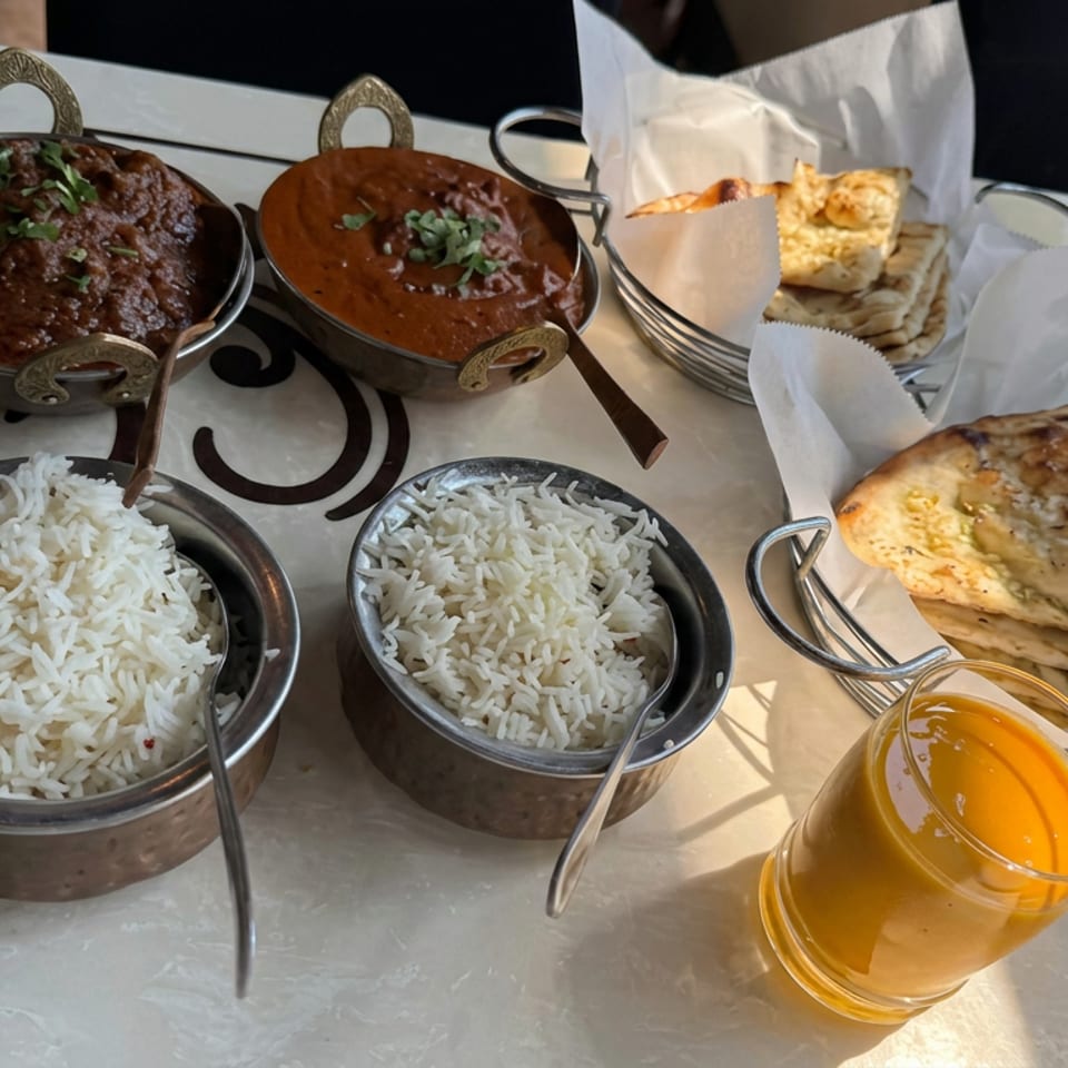 Indian meal featuring two curries, bowls of basmati rice, naan bread, and a glass of mango drink on a restaurant table.