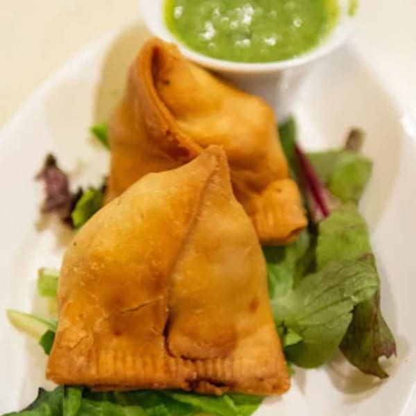 Two golden-brown Indian samosas on a bed of fresh greens, with a side of green chutney.