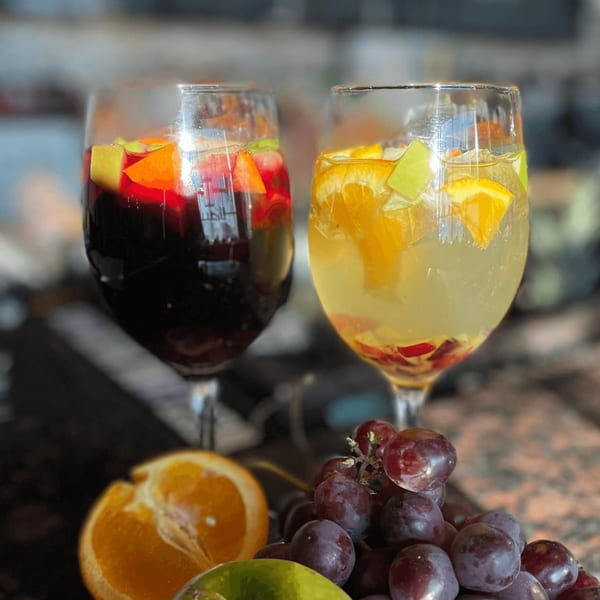 Two glasses of sangria, one red and one white, with fresh fruit, next to grapes and an orange.