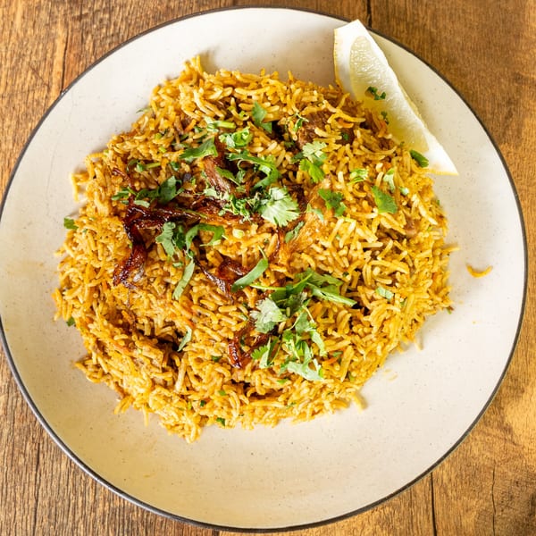 Indian biryani with rice, meat, cilantro, fried onions, and a lemon wedge on a plate.
