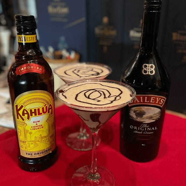 Two espresso martinis with chocolate drizzle, Kahlua, and Baileys bottles on a red surface.